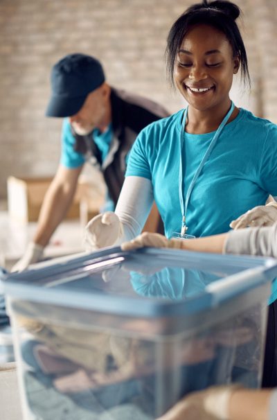 Group of people volunteering at donation center and packing clothes into boxes. Focus is on happy African American woman.